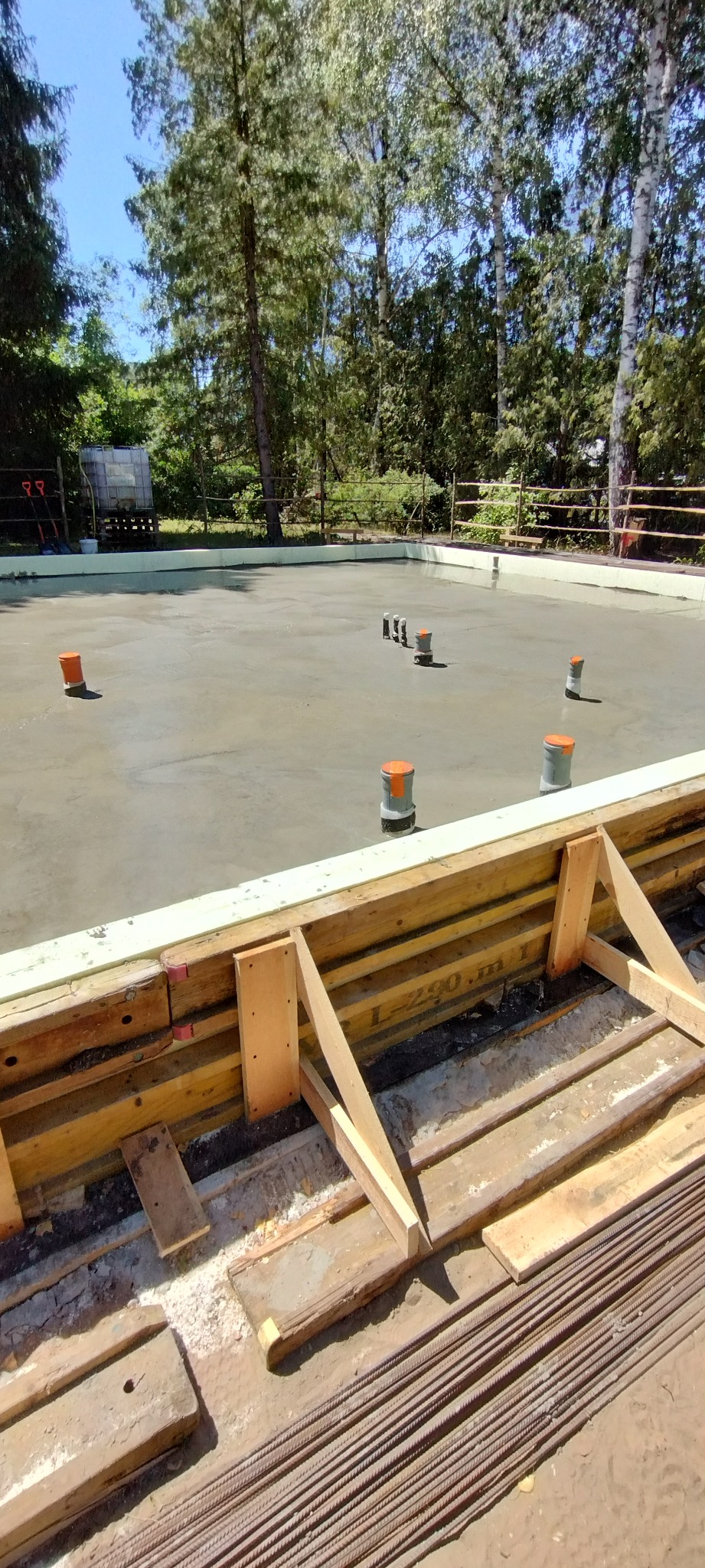 Świeżo wylany fundament pod budowę, z widocznym szalunkiem i wystającymi rurami wentylacyjnymi. Na pierwszym planie leżą pręty zbrojeniowe.