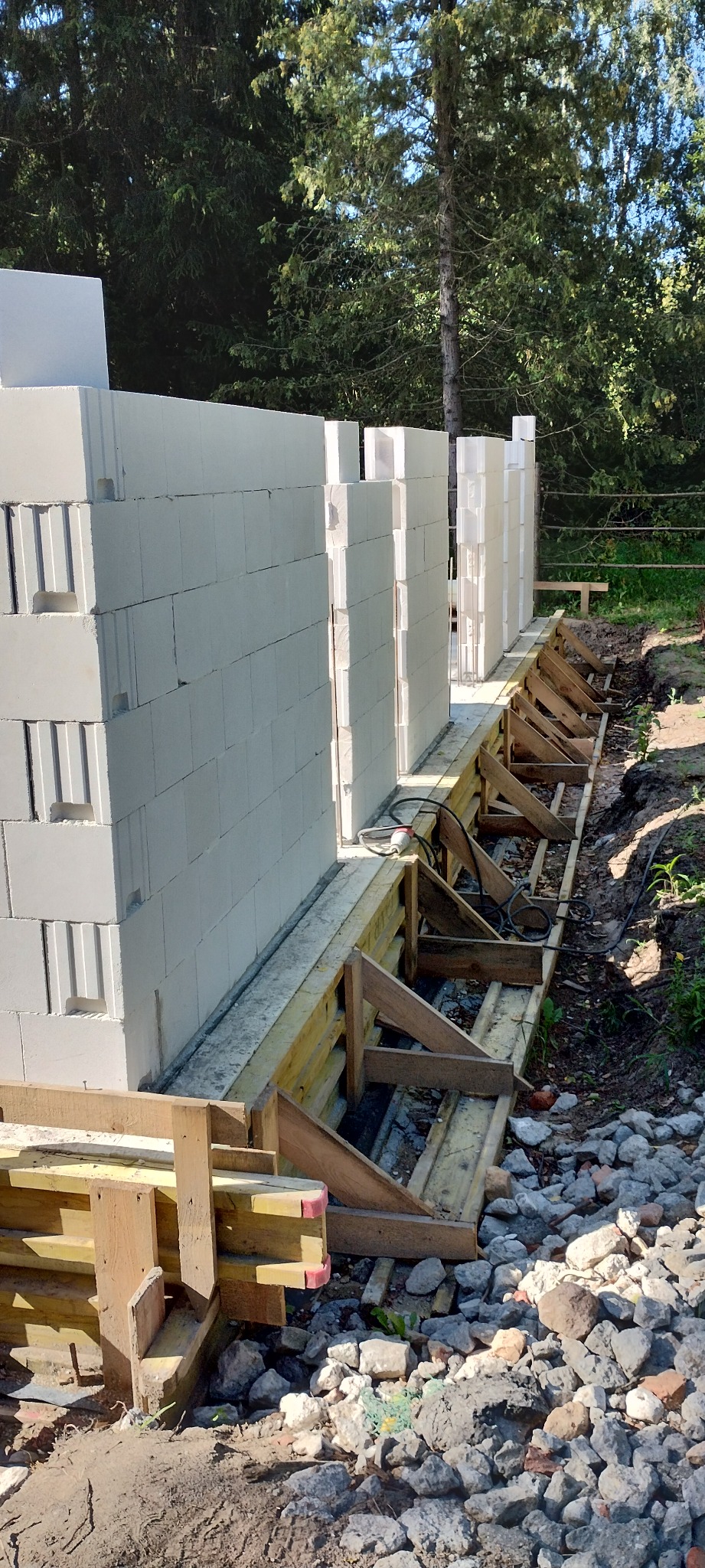 Budowa domu z bloczków betonu komórkowego. Widoczne szalunki fundamentowe i początek wznoszenia ścian. W tle drzewa.