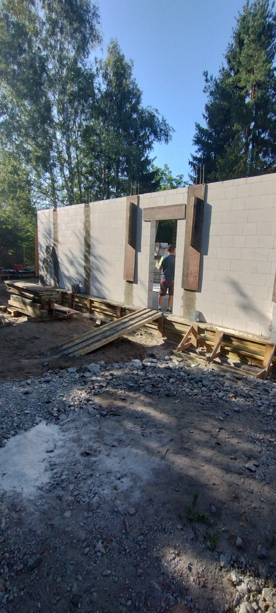 Wznoszenie ściany z betonu komórkowego Ytong. Widoczne drewniane szalunki okienne i zbrojenie. Prace budowlane w toku, teren budowy z gruzem i deskami.