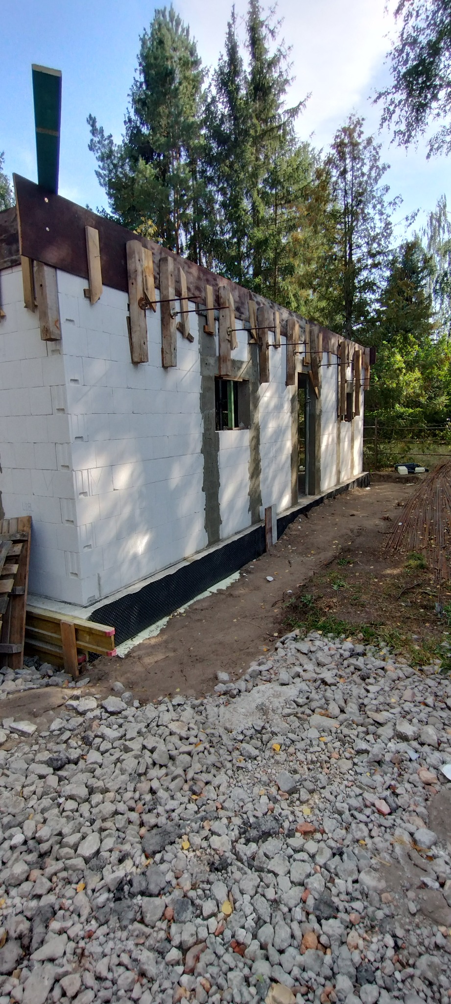 Budowa domu z białych bloczków, widoczne drewniane szalunki i izolacja fundamentów. W tle drzewa i niebo. Częściowo ułożone zbrojenie.