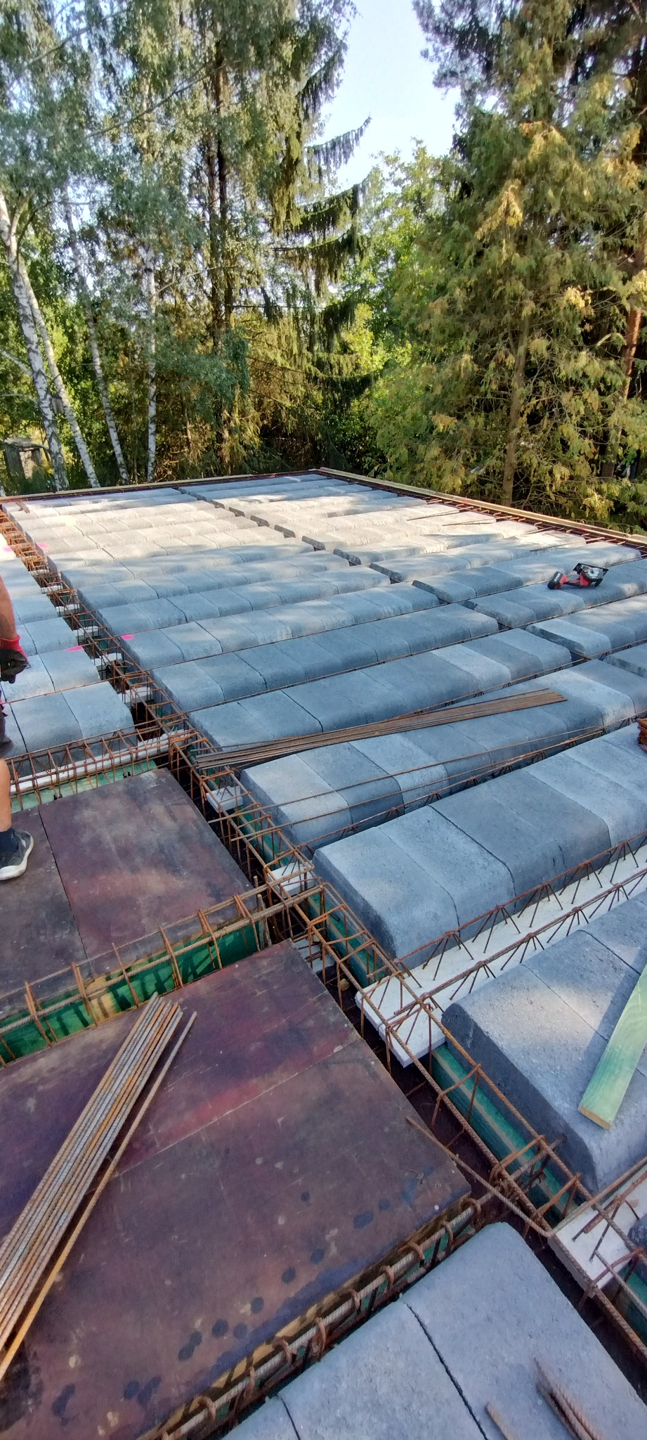 Zbrojenie stropu Teriva z betonowymi pustakami, widoczne pręty zbrojeniowe i deski szalunkowe. W tle drzewa.