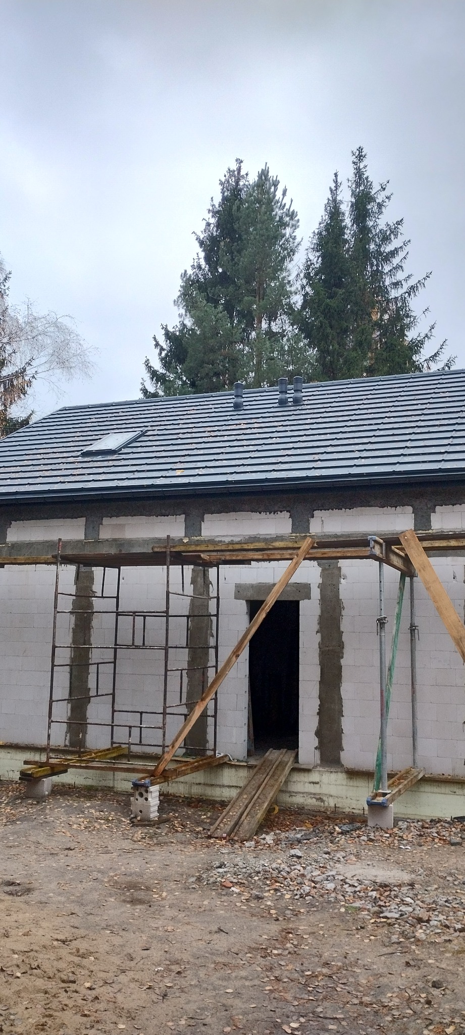 Budowa domu z białych bloczków, widoczne rusztowania i fragment dachu z dachówką. Surowy stan, jesienna sceneria z opadłymi liśćmi. Ujęcie z dołu, pochmurne niebo.