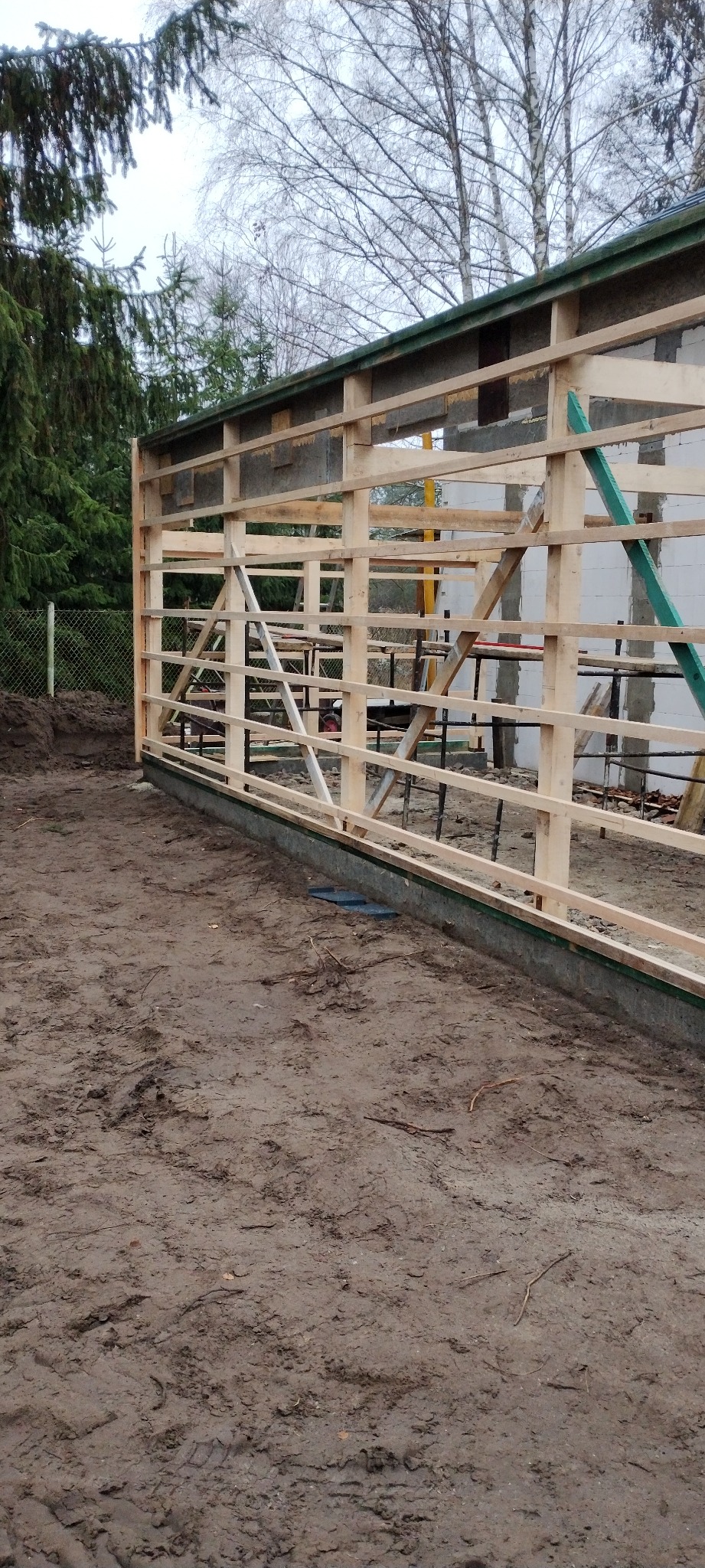 Konstrukcja szkieletowa domu w trakcie budowy. Widoczne drewniane elementy ramy na fundamencie, teren budowy z ziemią. W tle drzewa i częściowo ściana z bloczków.