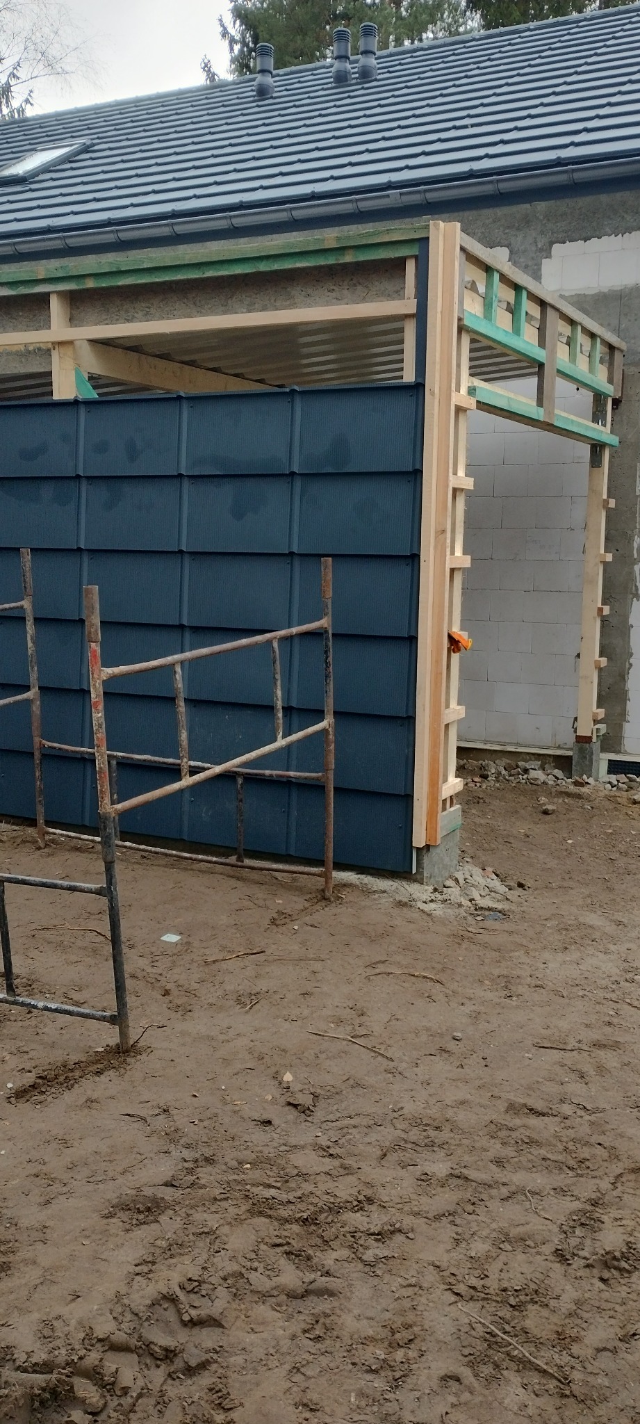 Budowa wiaty z drewnianą konstrukcją i ciemnymi panelami. Widoczne rusztowanie i fragment domu z bloczków. Prace w toku, teren wokół niezagospodarowany.