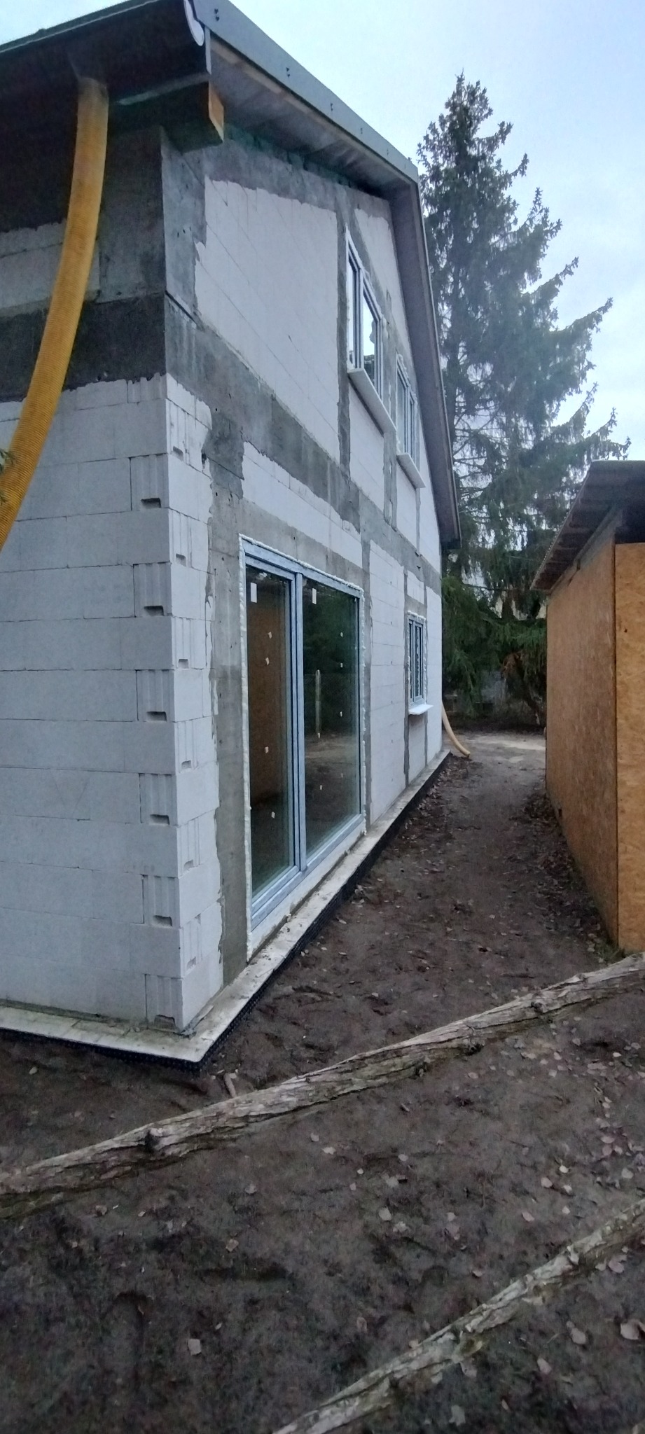 Budowa domu z betonu komórkowego: ściany w trakcie wznoszenia, widoczne okna i drzwi balkonowe. Na zewnątrz rura wentylacyjna i teren budowy.