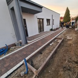 PAWEŁ MADEJ PKM BRUK - Nowoczesny dom z wybrukowanym chodnikiem z czerwono-brązowej kostki z białymi obrzeżami; widoczne przygotowania do ułożenia kostki w obramowaniu rabaty, niebieskie rury drenażowe i pracownicy w tle.