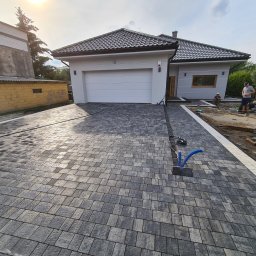 PAWEŁ MADEJ PKM BRUK - Nowoczesny dom jednorodzinny z podjazdem wyłożonym szarą kostką brukową w trakcie układania, widoczne niebieskie rury i pracownicy w tle.