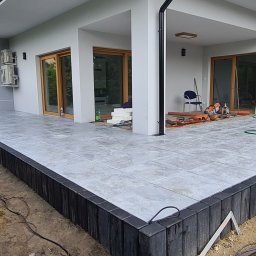 PAWEŁ MADEJ PKM BRUK - Wykończony taras z szarych płytek ceramicznych, obrzeża z czarnych palisad, fragment budynku z przeszklonymi drzwiami przesuwnymi i widocznymi narzędziami budowlanymi w tle.