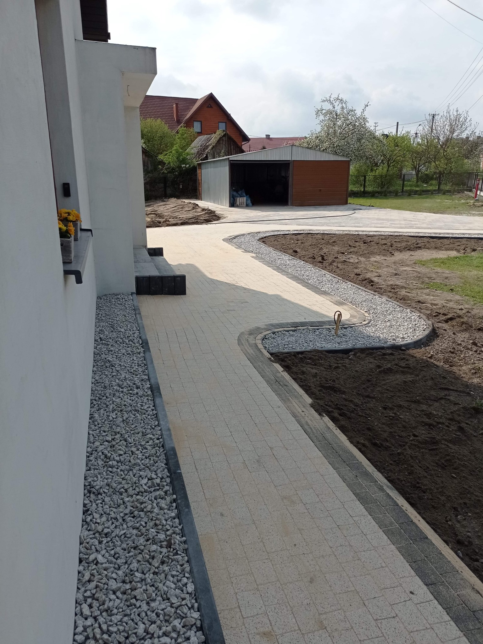 Ułożona ścieżka z jasnej kostki brukowej prowadząca od domu do garażu, obrzeża z ciemnej kostki i białego grysu, rabaty ziemne po bokach, widok z boku domu.