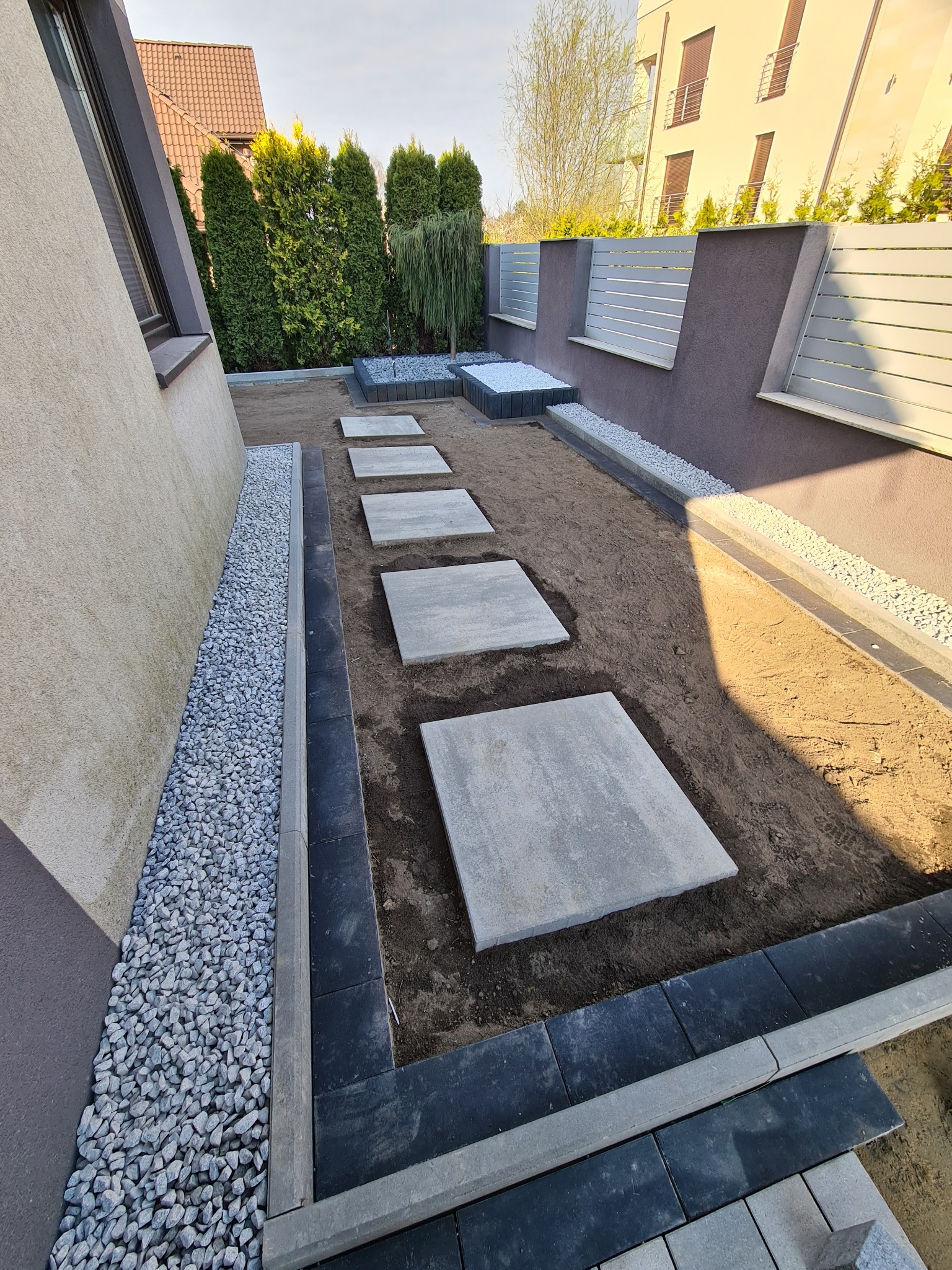 Ścieżka z kwadratowych płyt betonowych ułożona w ogrodzie, obrzeża wykończone kostką, po bokach dekoracyjny żwir, widok na nowoczesny dom i ogrodzenie.