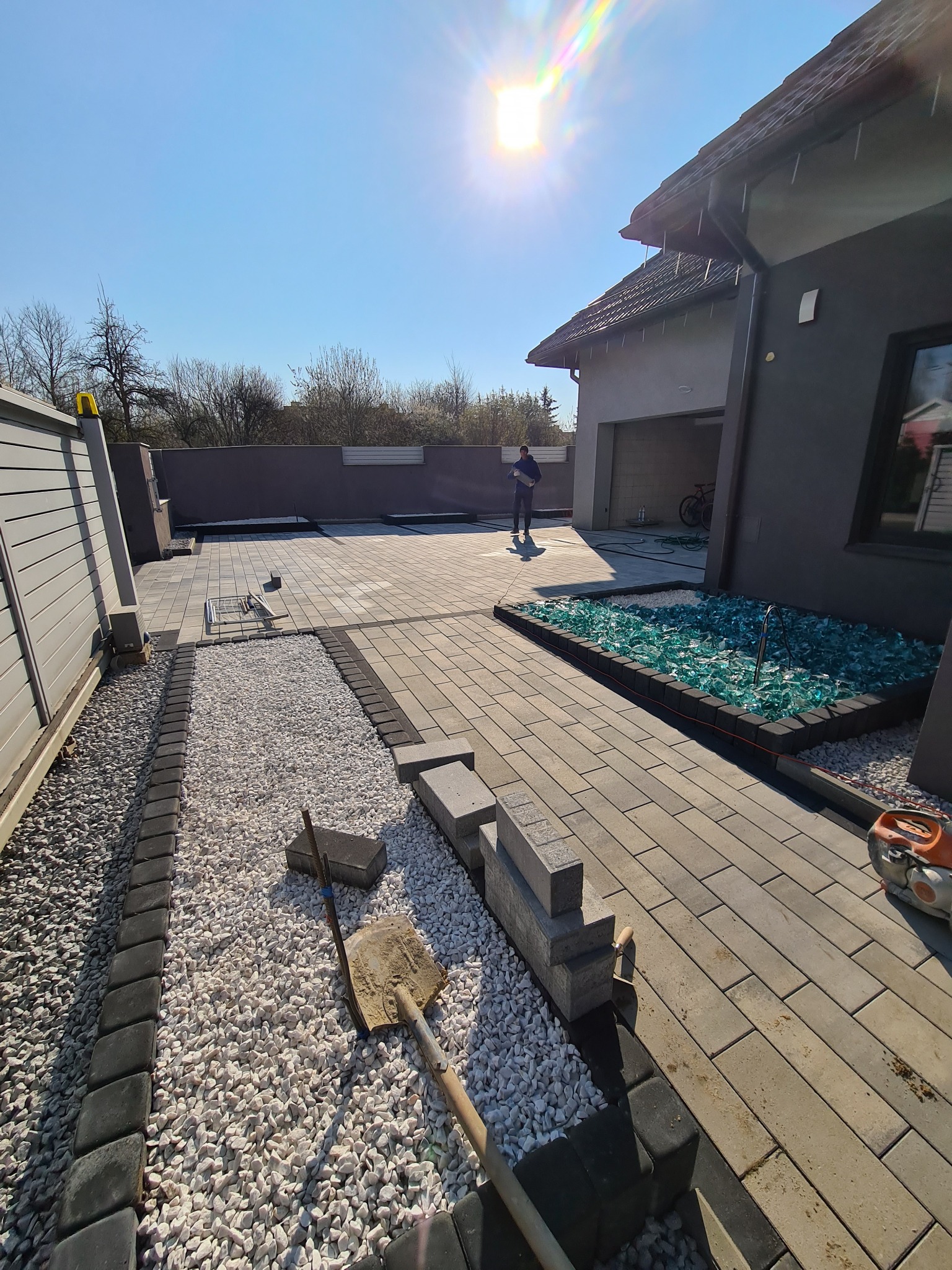 Nowoczesna posesja z jasnoszarą kostką brukową ułożoną w prostokątny wzór, obrzeża z ciemnej kostki, rabata z białym grysem i dekoracyjnym szkłem, widoczny mężczyzna i narzędzia brukarskie.