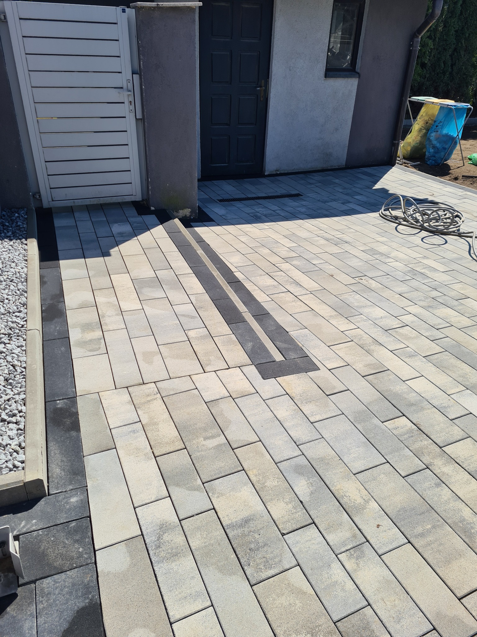 Ułożona kostka brukowa w odcieniach szarości z liniowym odpływem wody, widok z góry na posesji z białymi drzwiami i workami na śmieci w tle.