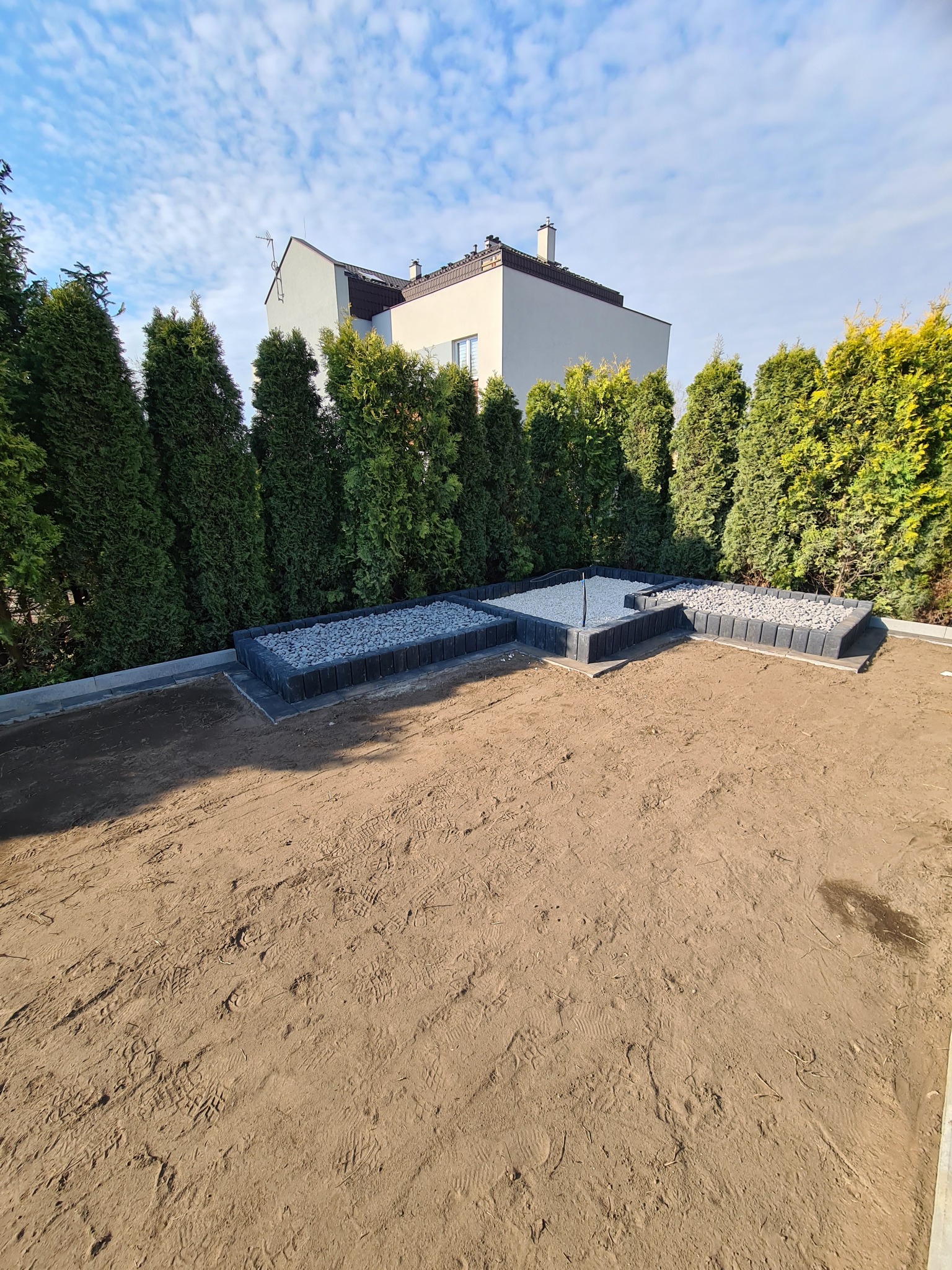 Przygotowany teren pod przyszły ogród z wyznaczonymi rabatami z obrzeżami wypełnionymi szarym grysem, tło: rząd tuj i budynek mieszkalny pod błękitnym niebem.