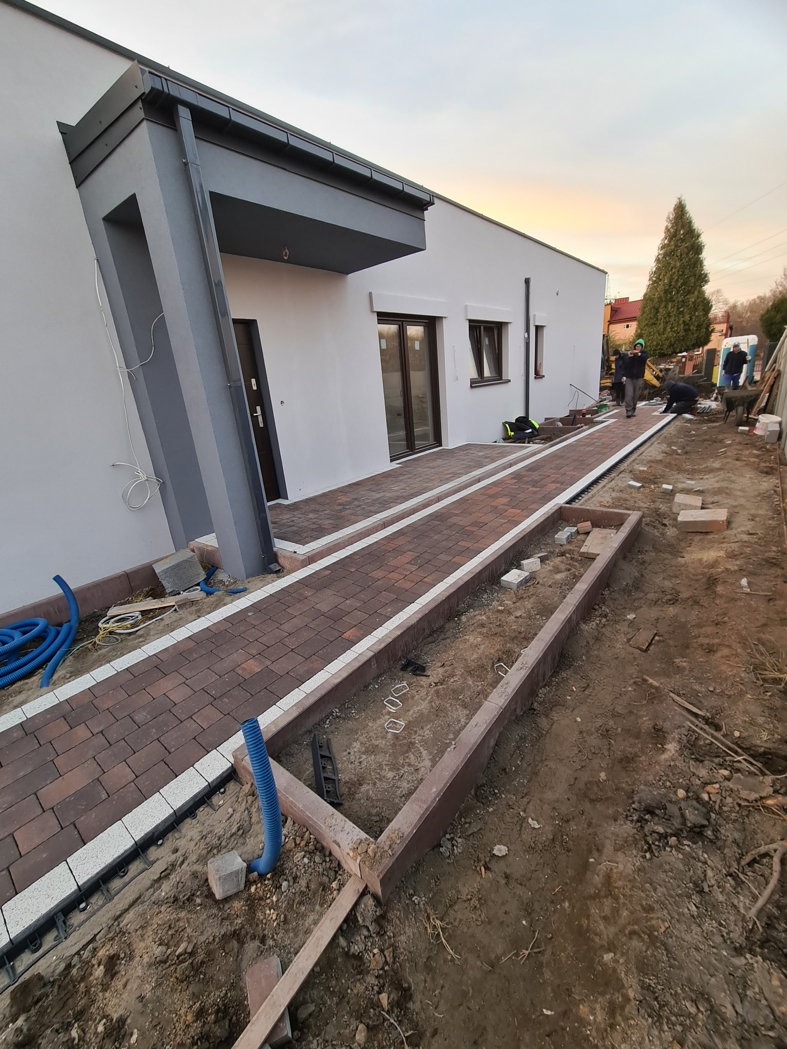 Nowoczesny dom z wybrukowanym chodnikiem z czerwono-brązowej kostki z białymi obrzeżami; widoczne przygotowania do ułożenia kostki w obramowaniu rabaty, niebieskie rury drenażowe i pracownicy w tle.