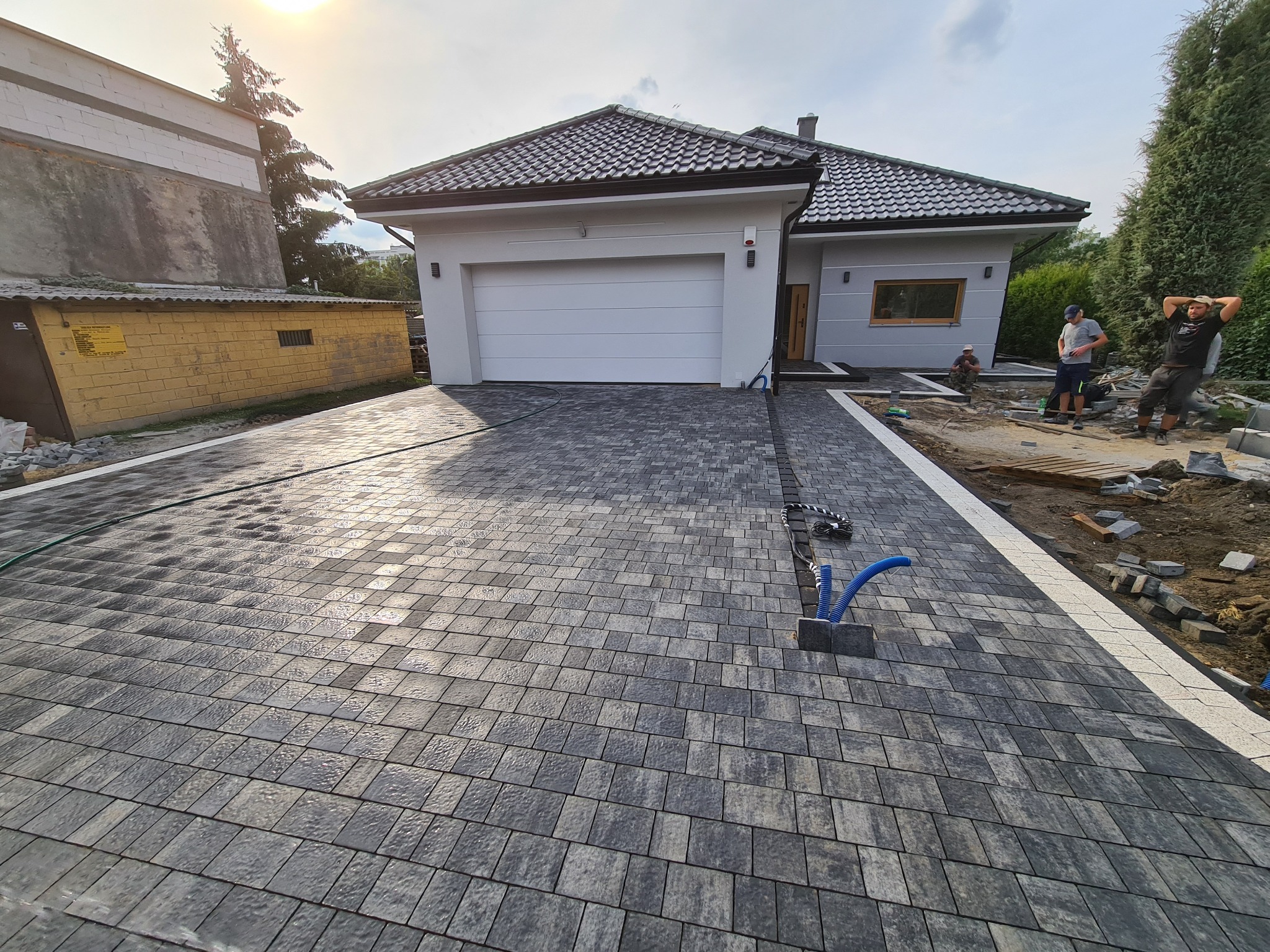 Nowoczesny dom jednorodzinny z podjazdem wyłożonym szarą kostką brukową w trakcie układania, widoczne niebieskie rury i pracownicy w tle.