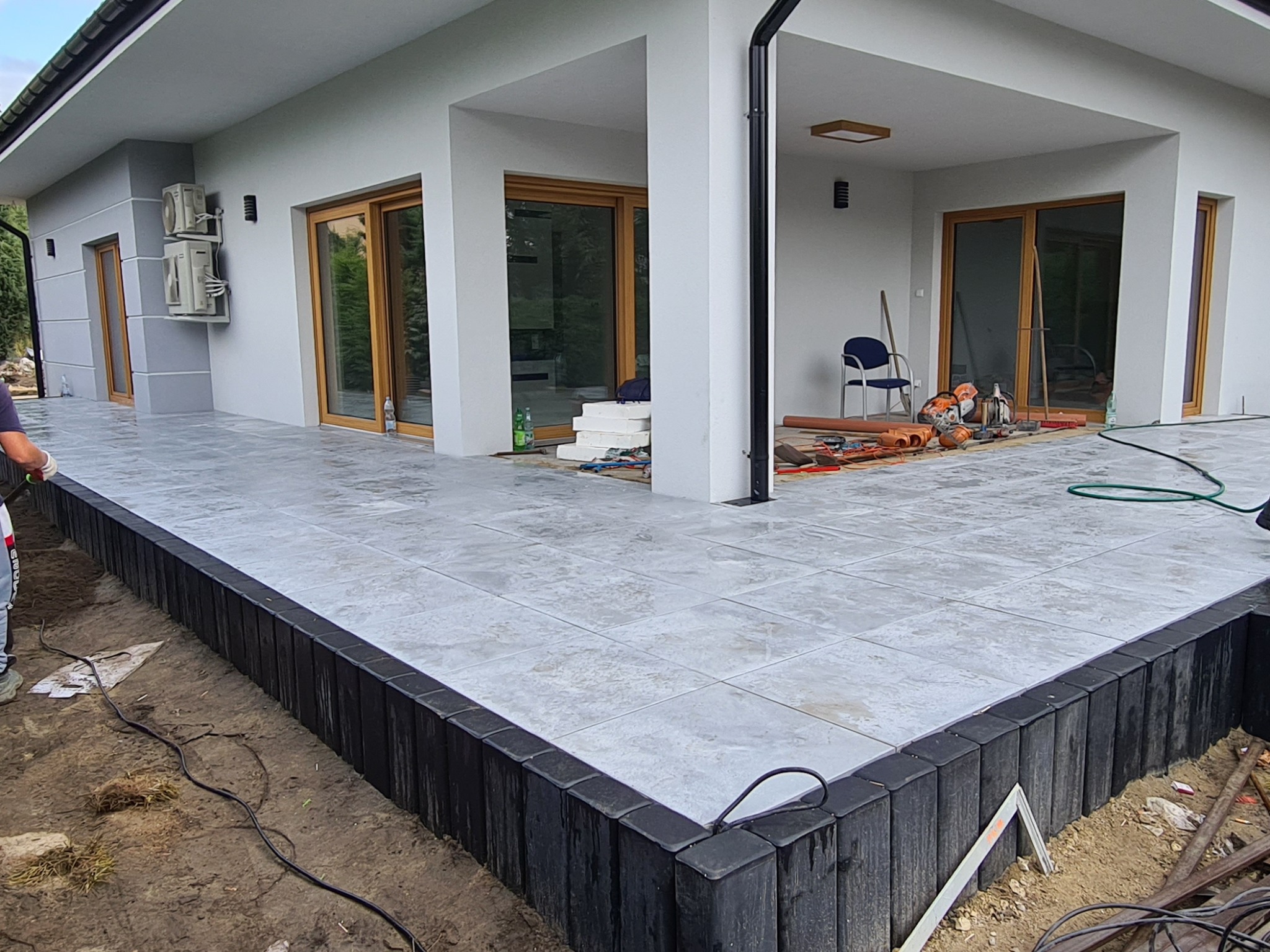 Wykończony taras z szarych płytek ceramicznych, obrzeża z czarnych palisad, fragment budynku z przeszklonymi drzwiami przesuwnymi i widocznymi narzędziami budowlanymi w tle.