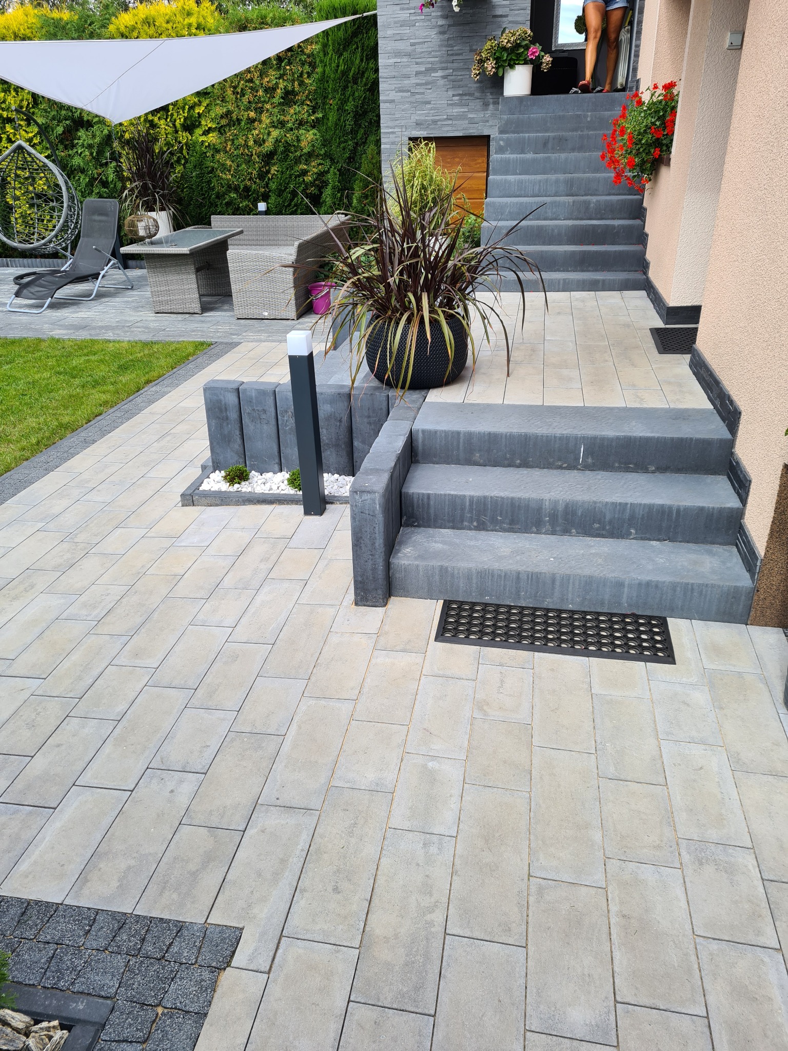 Nowoczesny taras z jasnej kostki brukowej w odcieniach beżu i szarości, prowadzący do schodów wejściowych z donicami kwiatów, oświetlony lampami ogrodowymi, z widocznym fragmentem strefy relaksu...