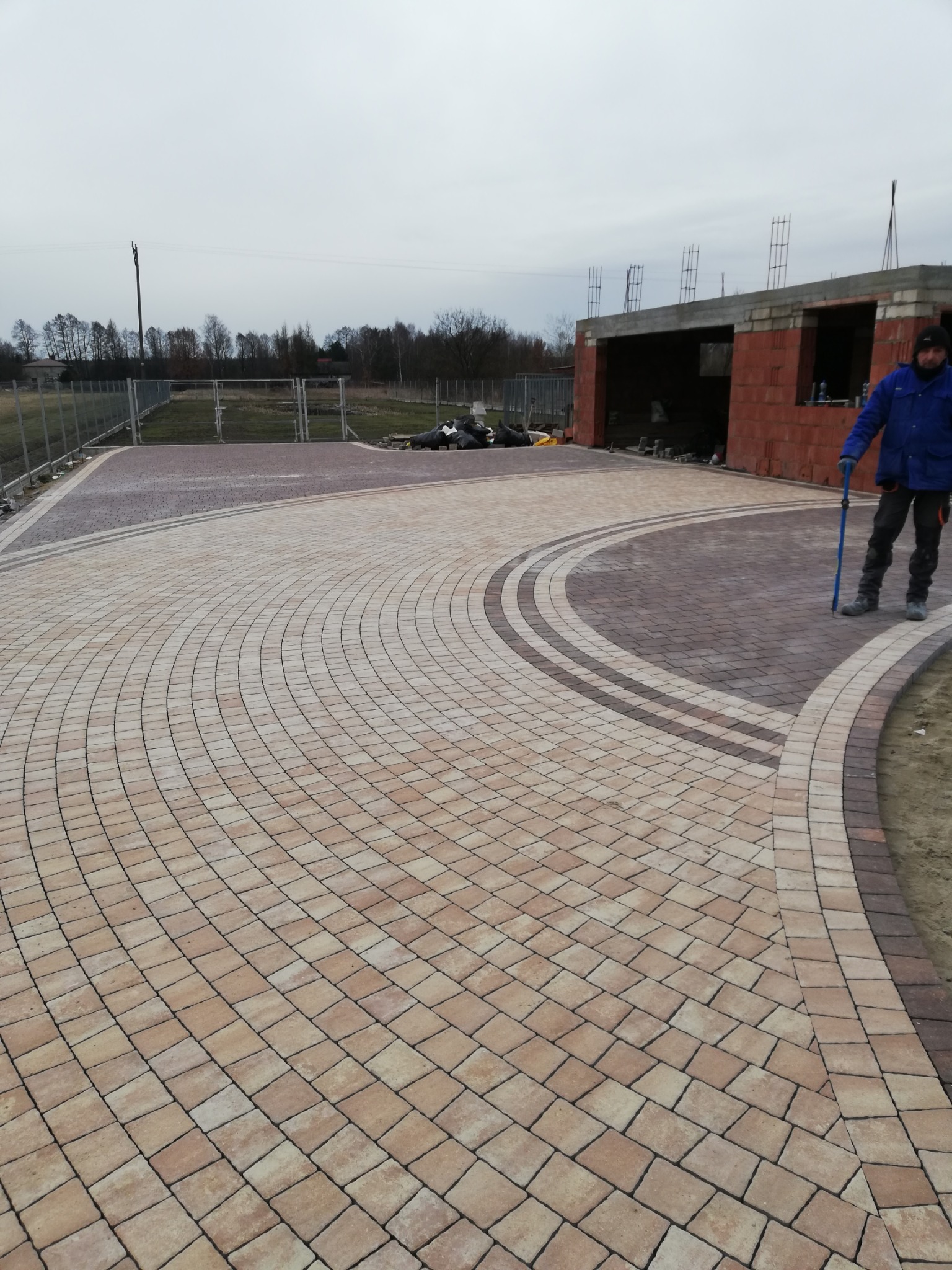 Ułożona kostka brukowa w odcieniach beżu i brązu, tworząca wzór łuków na podjeździe; w tle niedokończony budynek gospodarczy z czerwonej cegły i pracownik z niebieskim narzędziem.