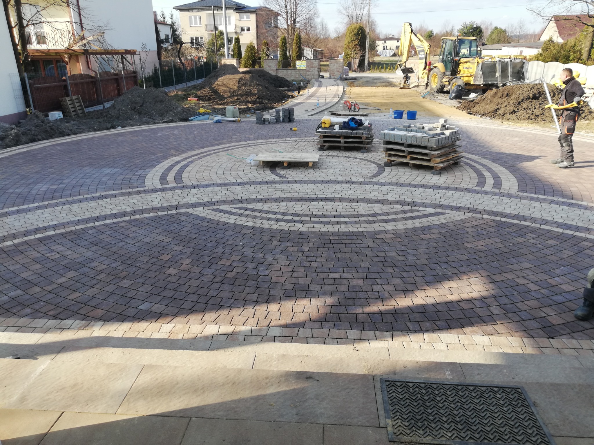 Okrągły podjazd z kostki brukowej ułożonej we wzór koncentrycznych okręgów, z paletami kostki, koparką w tle i pracownikiem w odzieży roboczej.