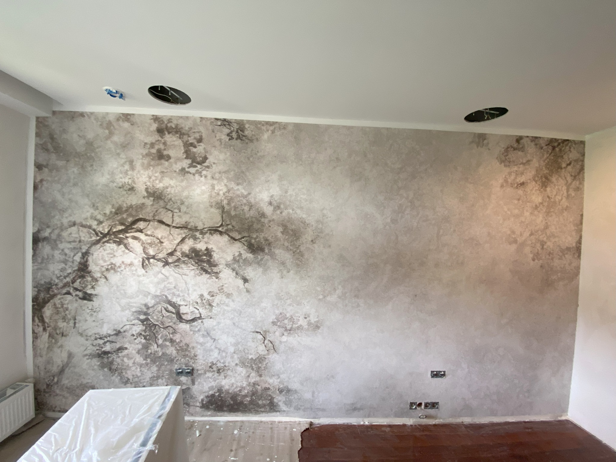 Ściana w pokoju pokryta dekoracyjną farbą w odcieniach szarości, przypominającą motyw drzewa, widoczne gniazdka elektryczne i fragment parkietu na podłodze, oraz otwory w suficie na oświetlenie.