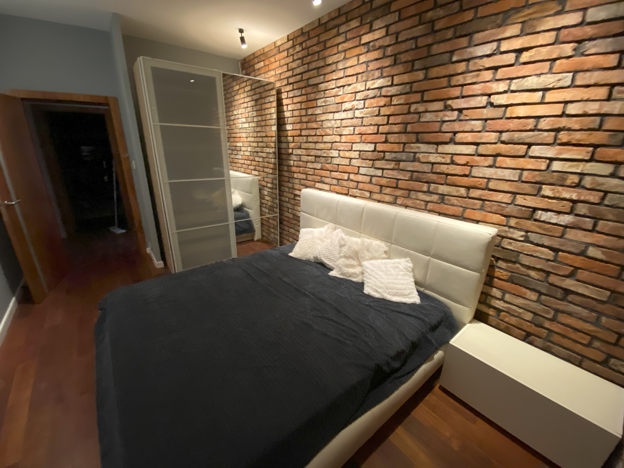 Sypialnia z łóżkiem z czarną narzutą i białymi poduszkami, ściana z czerwonej cegły, parkiet na podłodze, biała szafa z lustrzanymi drzwiami.