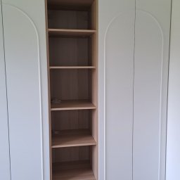 STOLARSTWO PAWEŁ KOPACZEWSKI - Biała szafa z półkami z jasnego drewna, widoczne pionowe frezy na górze szafy, podłoga z parkietu ułożonego we wzór jodełki.