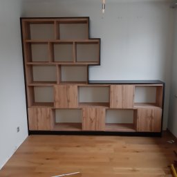 STOLARSTWO PAWEŁ KOPACZEWSKI - Narożna, drewniana biblioteczka z czarnymi obramowaniami, z otwartymi półkami i częściowo zabudowanymi szafkami, na tle białej ściany i drewnianej podłogi, z narzędziami widocznymi w rogu.