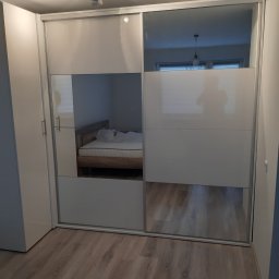 STOLARSTWO PAWEŁ KOPACZEWSKI - Biała szafa przesuwna z lustrzanymi drzwiami, widoczne odbicie łóżka w sypialni, oświetlenie sufitowe.