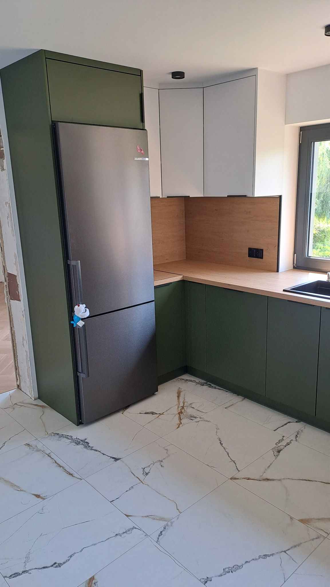 Nowoczesna kuchnia z oliwkowymi szafkami dolnymi, szarym lodówko-zamrażarką i białymi szafkami górnymi, wykończona drewnopodobnym blatem i panelem ściennym, z marmurową podłogą.