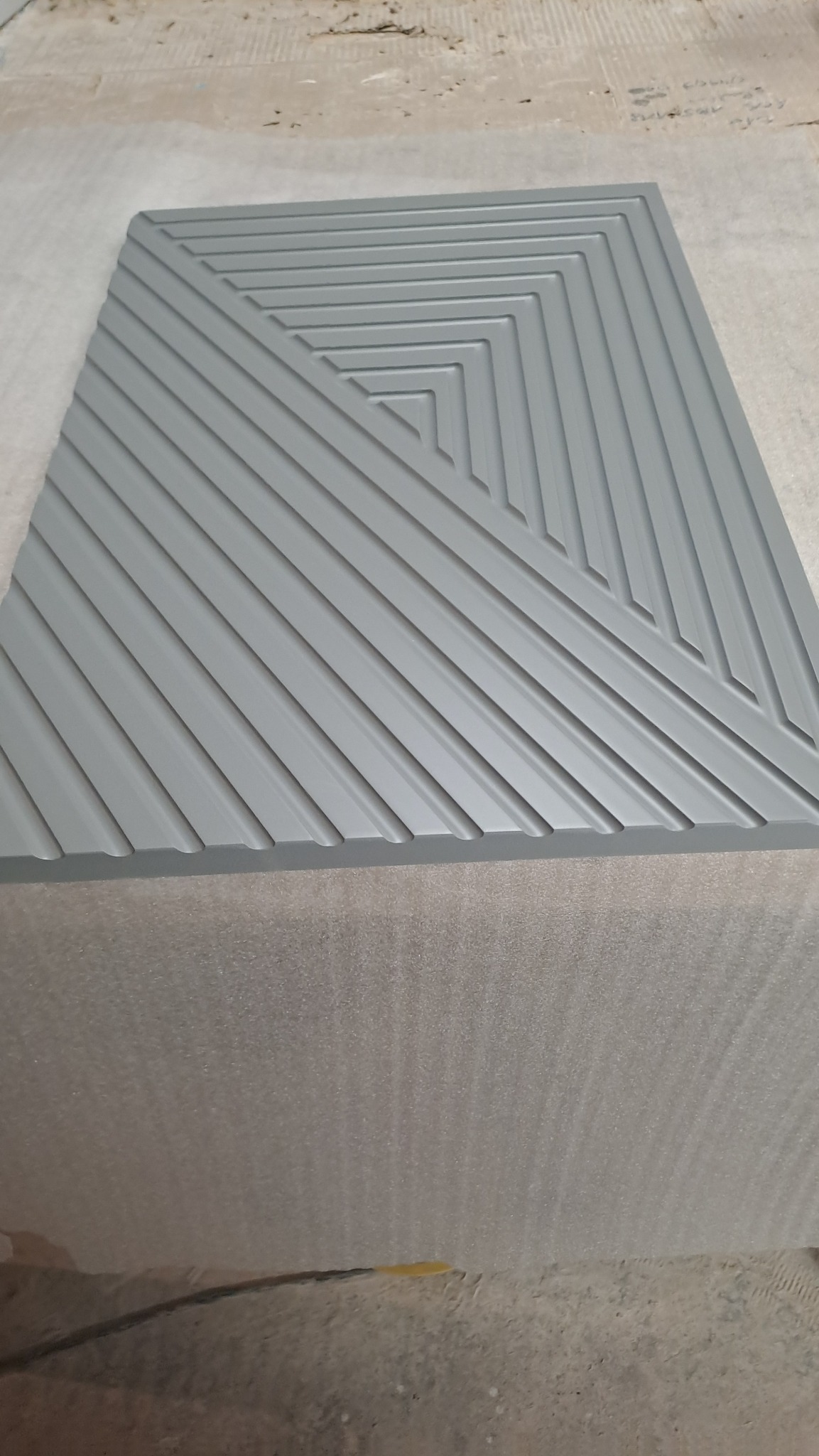 Szara płyta MDF z frezowanym geometrycznym wzorem koncentrycznych kwadratów, leżąca na podłożu z pianki.