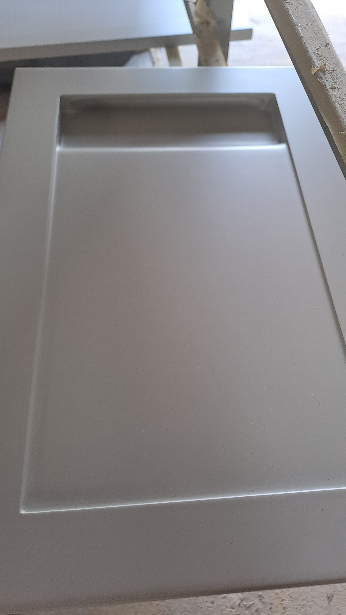 Szary, lakierowany front meblowy z frezowanym uchwytem, leżący na drewnianym podłożu.