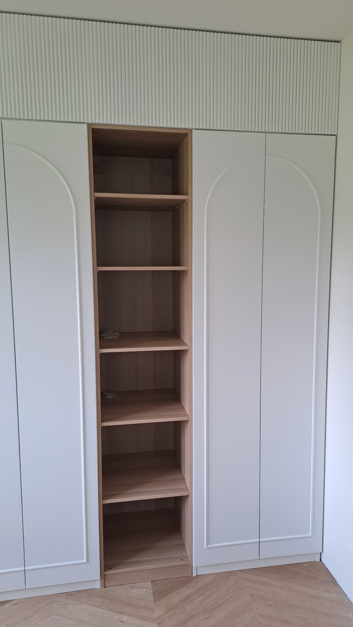 Biała szafa z półkami z jasnego drewna, widoczne pionowe frezy na górze szafy, podłoga z parkietu ułożonego we wzór jodełki.