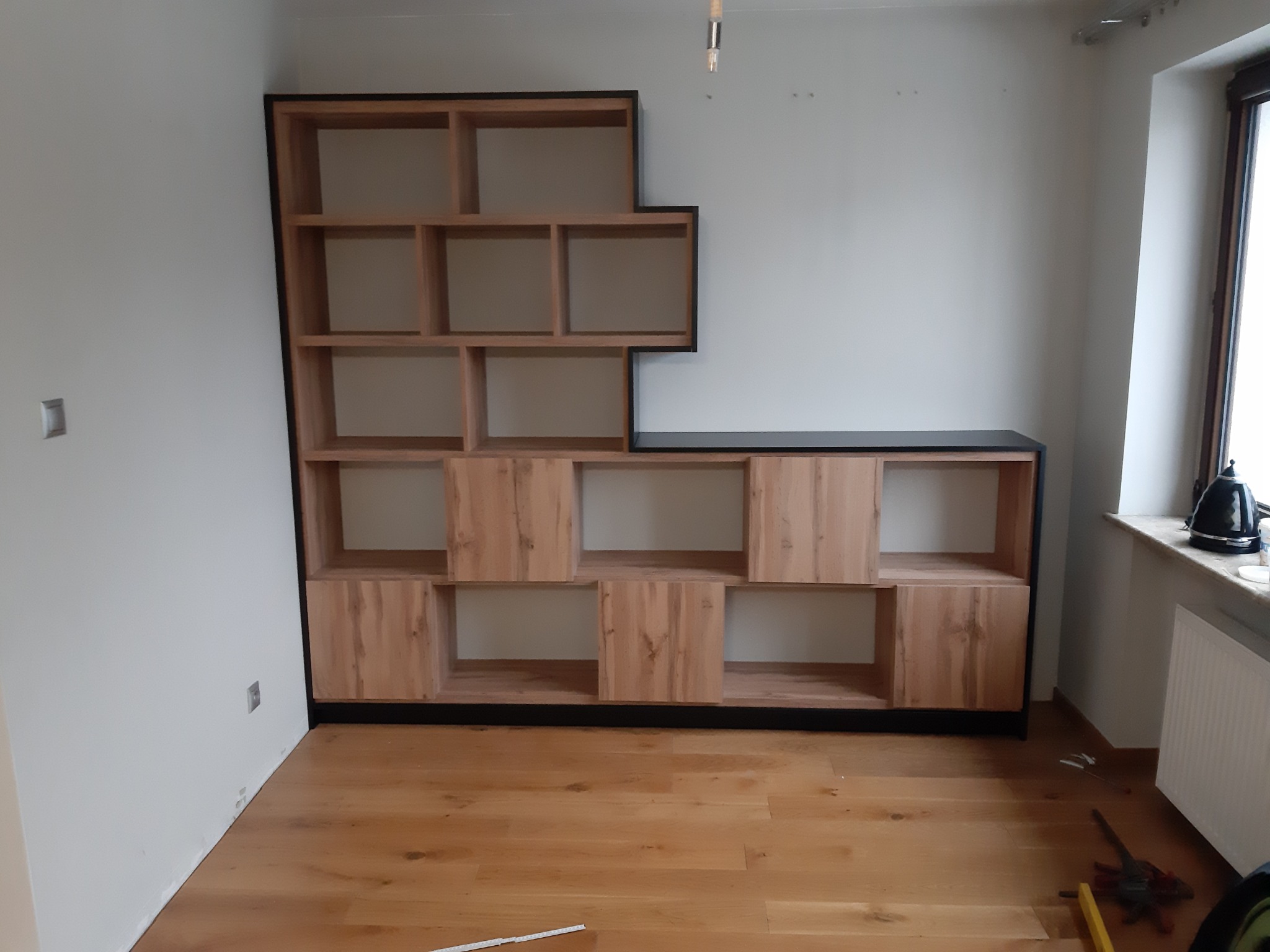 Narożna, drewniana biblioteczka z czarnymi obramowaniami, z otwartymi półkami i częściowo zabudowanymi szafkami, na tle białej ściany i drewnianej podłogi, z narzędziami widocznymi w rogu.