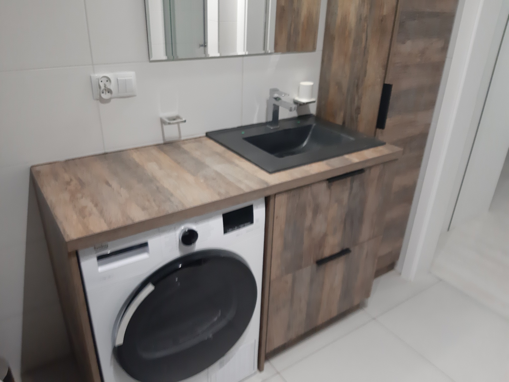 Łazienka z pralką zabudowaną pod blatem z umywalką i szafką w kolorze drewna, białe płytki na ścianach, minimalistyczny design.