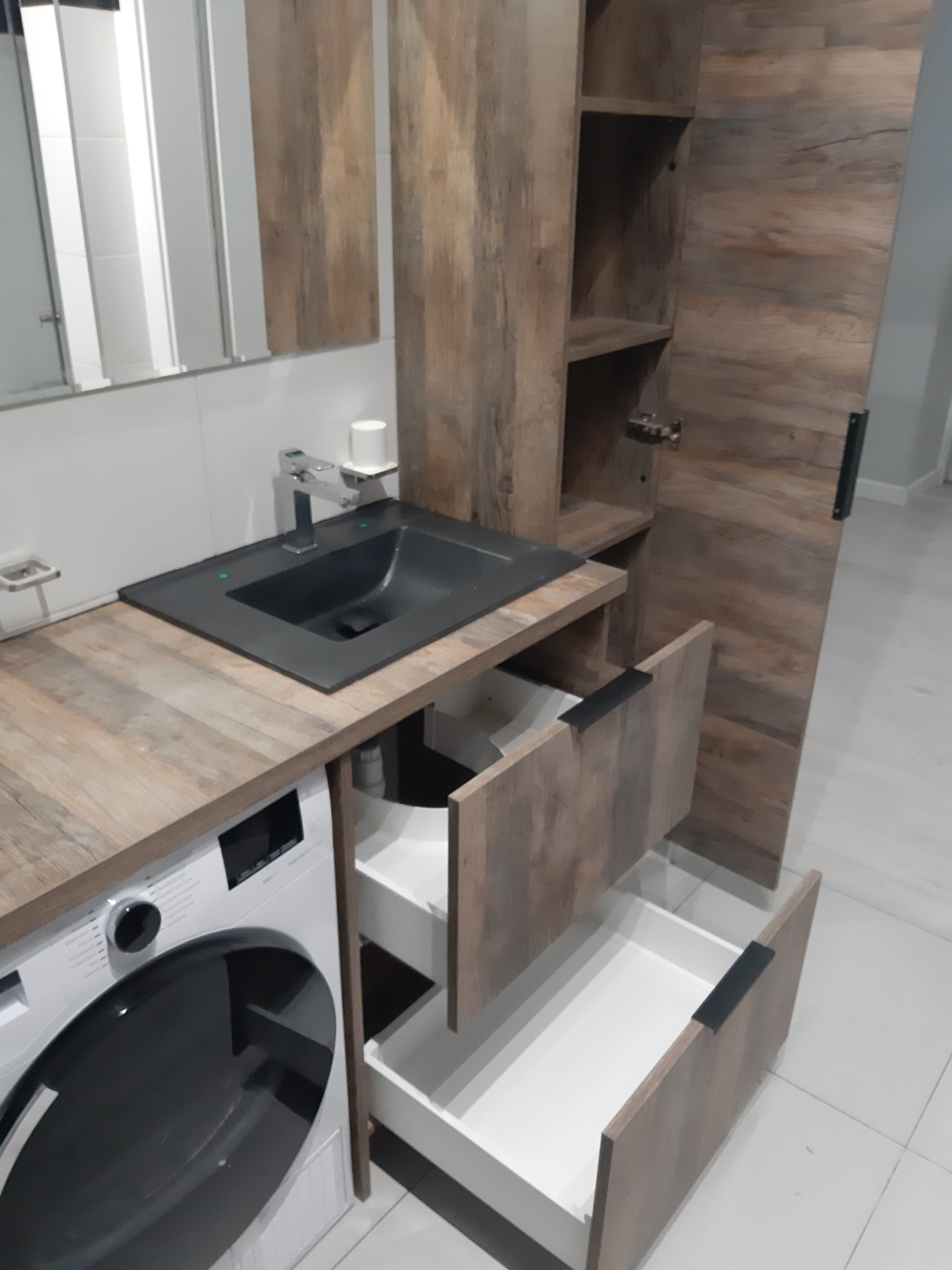 Łazienka z zabudową meblową: umywalka nablatowa w kolorze czarnym, drewnopodobny blat, pralka pod blatem, szafka z otwartymi szufladami i półkami.
