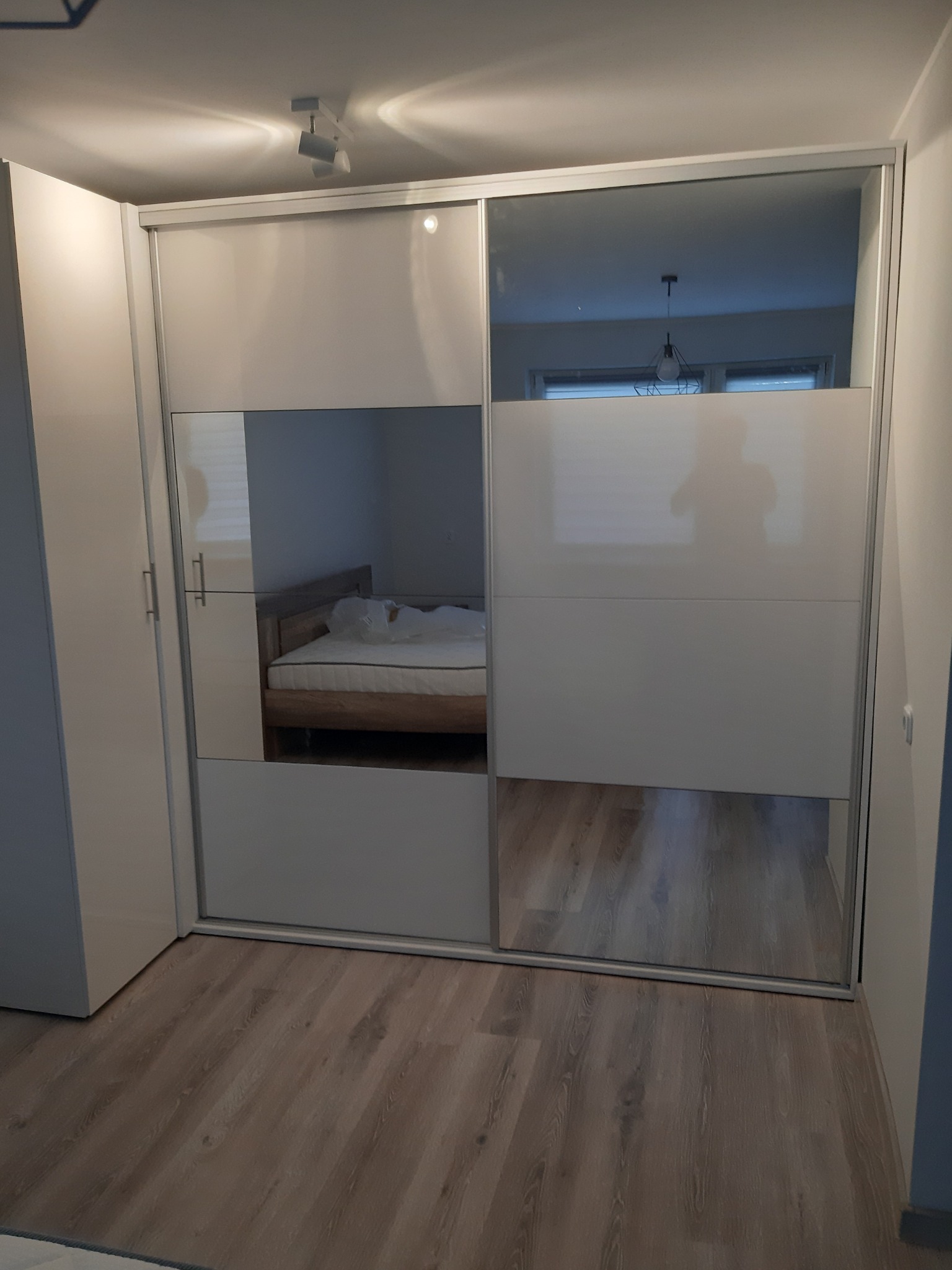 Biała szafa przesuwna z lustrzanymi drzwiami, widoczne odbicie łóżka w sypialni, oświetlenie sufitowe.