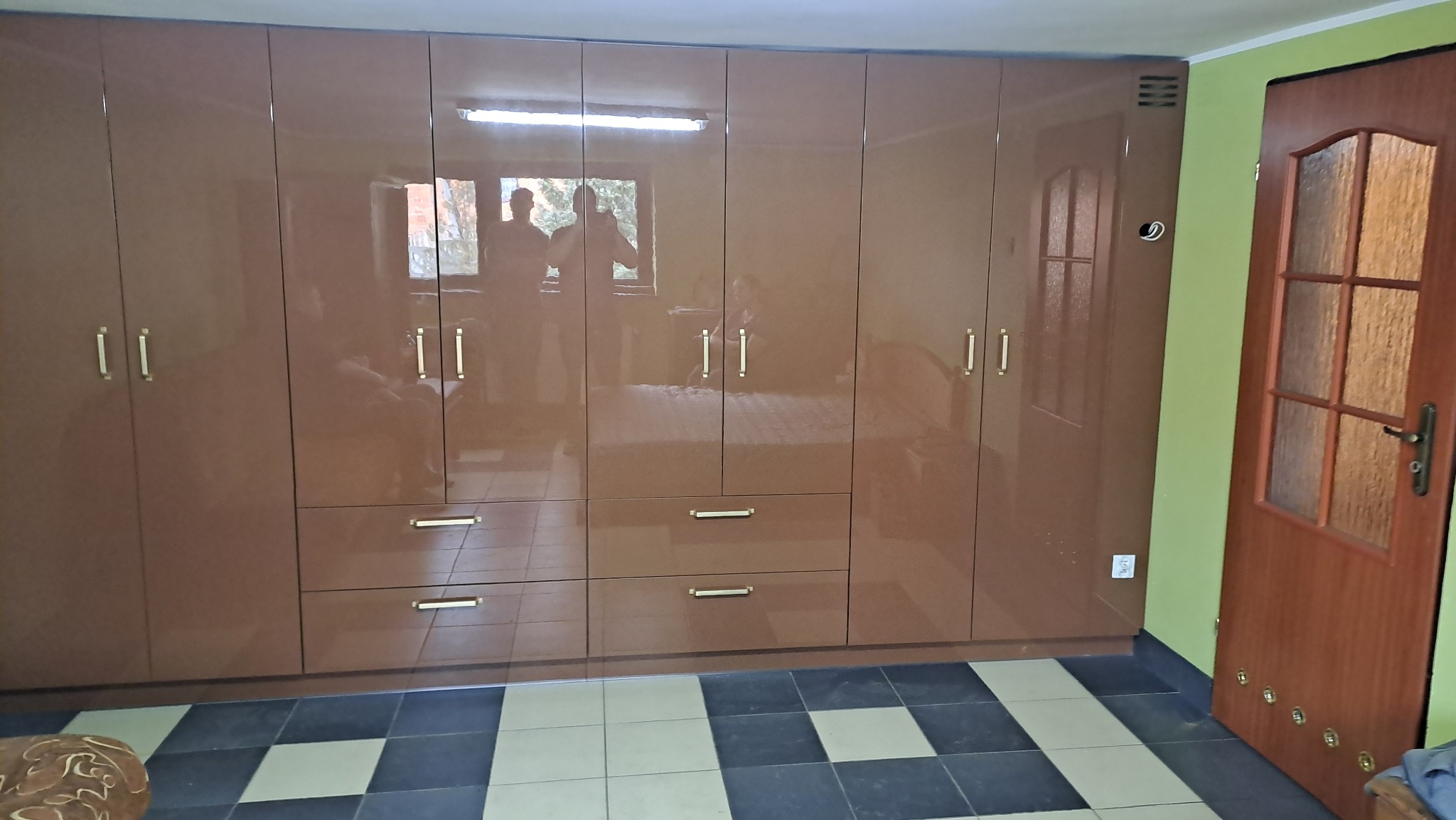 Duża, brązowa szafa wnękowa z połyskiem, z szufladami w dolnej części i złotymi uchwytami, odbijająca sylwetki osób w drzwiach, na podłodze czarno-biała szachownica.