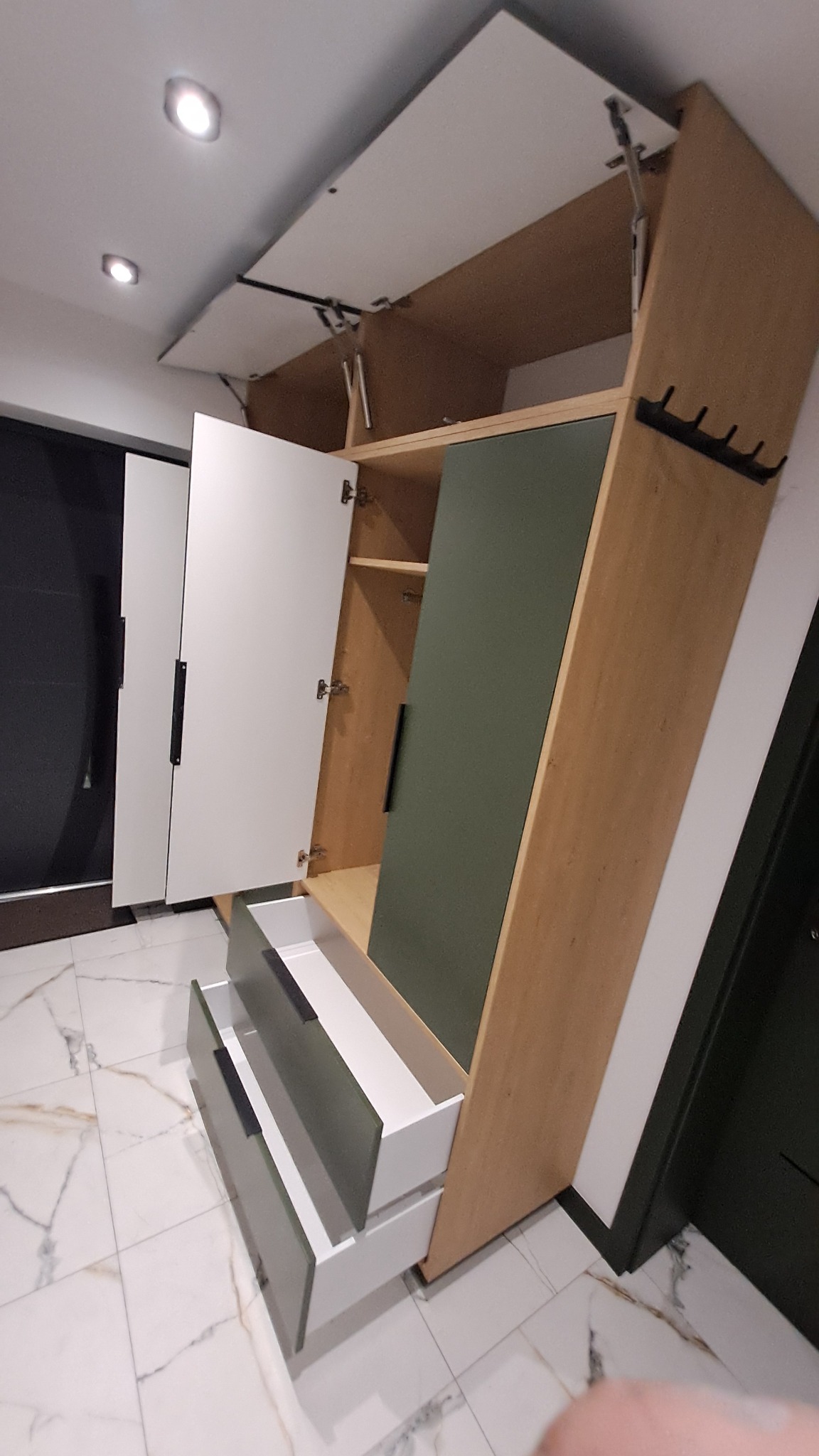 Otwarta szafa w przedpokoju z drewnianymi elementami, zielonym panelem i szufladami z szarymi frontami, widoczne mechanizmy podnoszące górną część szafy.