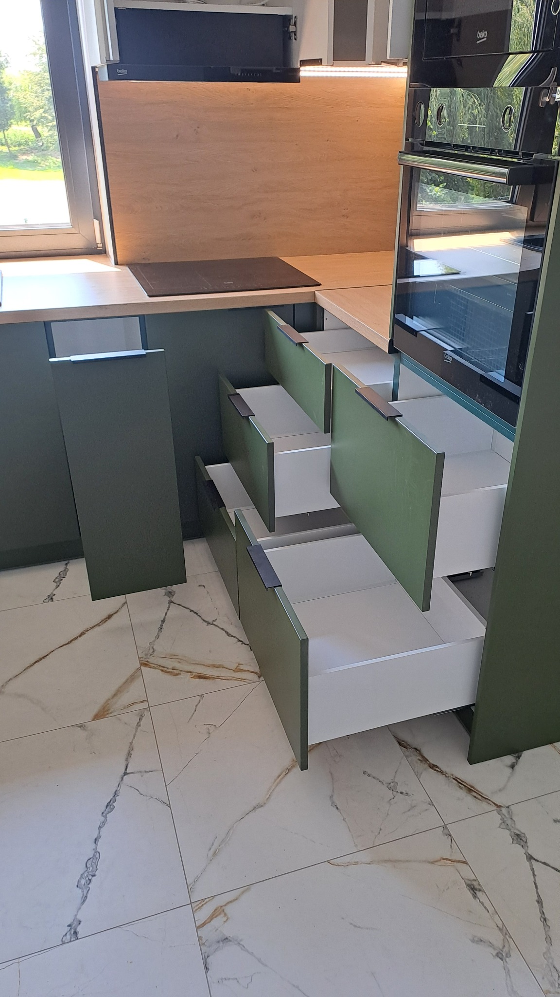 Nowoczesna kuchnia z zielonymi frontami i białymi wnętrzami szuflad, częściowo wysunięte szuflady dolne, widoczny piekarnik i płyta indukcyjna, marmurowa podłoga.