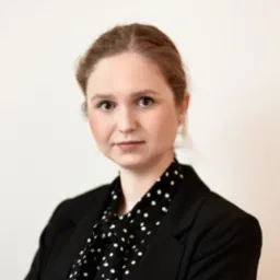 Portret kobiety w czarnym żakiecie i bluzce w białe kropki na jasnym tle.