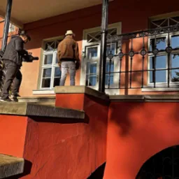 Ekipa filmowa kręci scenę teledysku na schodach budynku o czerwonej elewacji. Jeden mężczyzna filmuje kamerą, drugi stoi na podeście przed oknem.