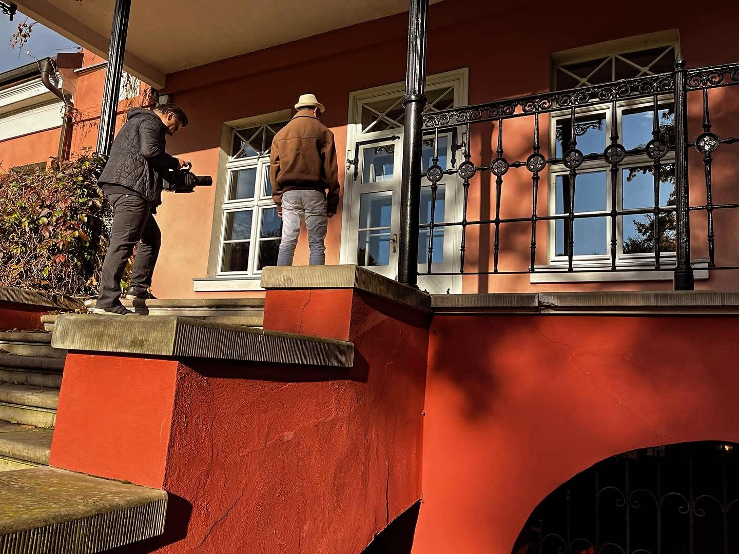 Ekipa filmowa kręci scenę teledysku na schodach budynku o czerwonej elewacji. Jeden mężczyzna filmuje kamerą, drugi stoi na podeście przed oknem.