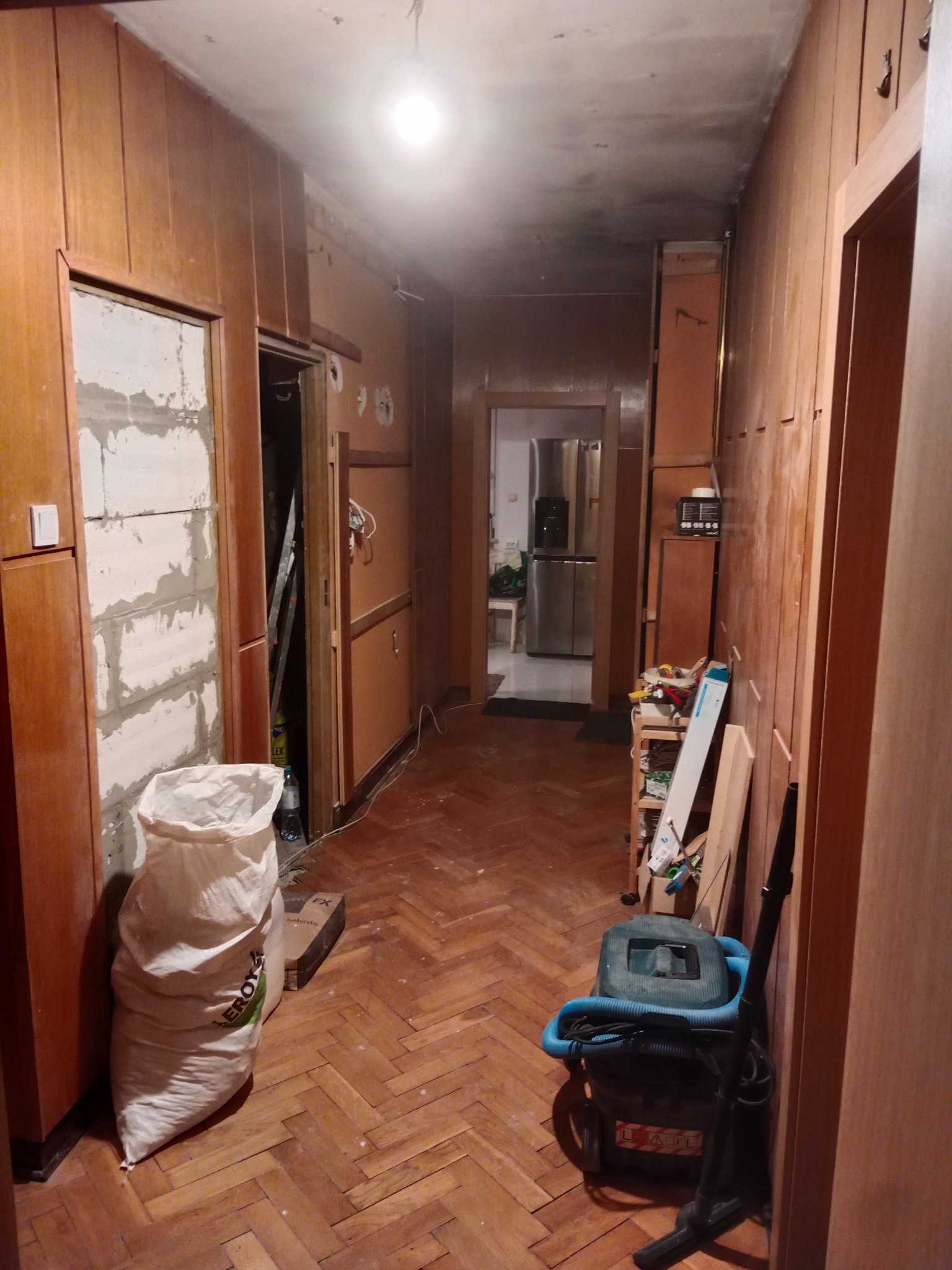 Korytarz w trakcie remontu: zamurowane drzwi, odsłonięte cegły, panele parkietowe w jodełkę, widoczny worek z zaprawą budowlaną Leroy Merlin oraz odkurzacz przemysłowy.