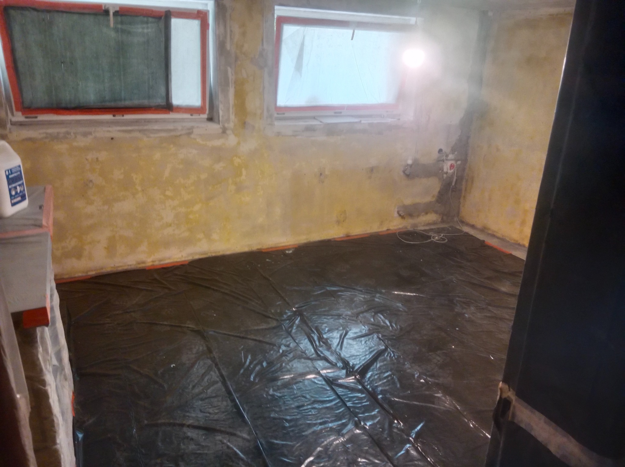 Przygotowanie pomieszczenia do prac wykończeniowych: zabezpieczona folią podłoga, okna z pomarańczową ramą zabezpieczone folią, ściany w trakcie gruntowania, widoczne instalacje elektryczne.