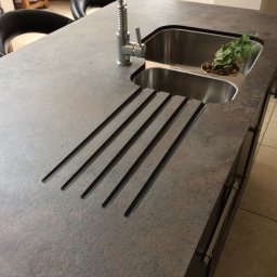 MJ Serwis - Blat kuchenny z szarego kamienia z wbudowanym zlewem, ociekaczem w postaci wyfrezowanych rowków i małą roślinką w doniczce.