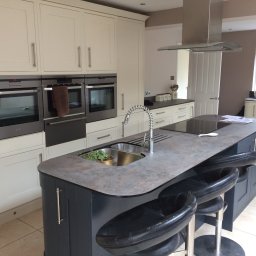 MJ Serwis - Jasna kuchnia z wyspą, szarym blatem i czarnymi hokerami, widoczne trzy piekarniki w zabudowie i roślina w złotej doniczce w zlewie.