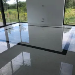 Błyszcząca, jasnoszara podłoga z płytek z czarnymi akcentami w nowocześnie wykończonym, jasnym pomieszczeniu z dużymi oknami i widokiem na zieleń na zewnątrz.