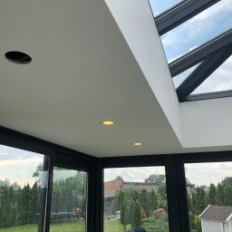 MJ Serwis - Wnętrze przeszklonej werandy z widokiem na ogród w deszczowy dzień, widoczne oczka oświetleniowe w suficie i otwór na montaż kolejnego punktu świetlnego.