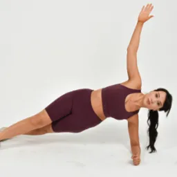 Kobieta w sportowym stroju w kolorze burgund wykonuje pozycję boczną deski na białym tle, z uniesioną prawą ręką i wzrokiem skierowanym w stronę kamery.