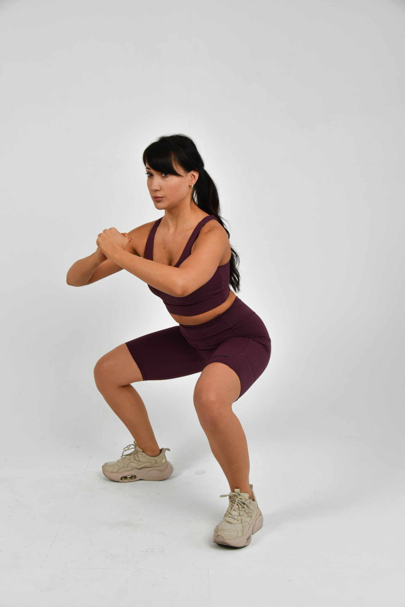 Kobieta w burgundowym stroju sportowym wykonuje przysiad ze złączonymi dłońmi na tle białego studia, prezentując ćwiczenie fitness.
